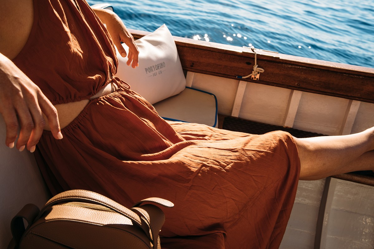 Woman in rust-colored dress seated in boat on sunny day with backpack, white cushion. Blue water visible in background.