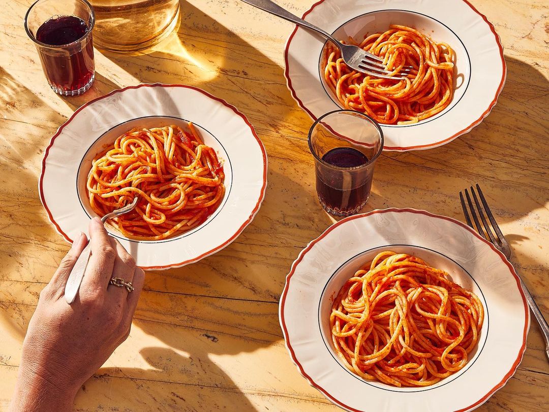 Overhead shot of three plates of spaghetti with tomato sauce, a fork in one plate, and drinks on a marble table, creating a vibrant food scene.