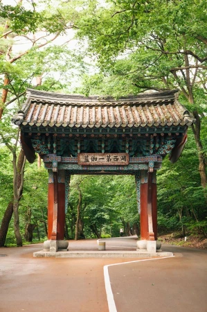 Ornate arched gate with a tiled roof stands in a lush, green park in Asia, welcoming visitors along a winding pathway.