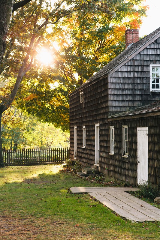 Exterior of cedar shake house with white trim and a red brick chimney on a sunny day with trees and grass visible.