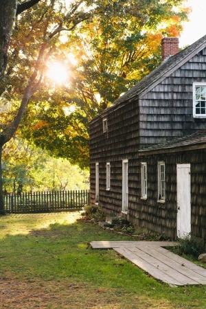 Exterior of cedar shake house with white trim and a red brick chimney on a sunny day with trees and grass visible.