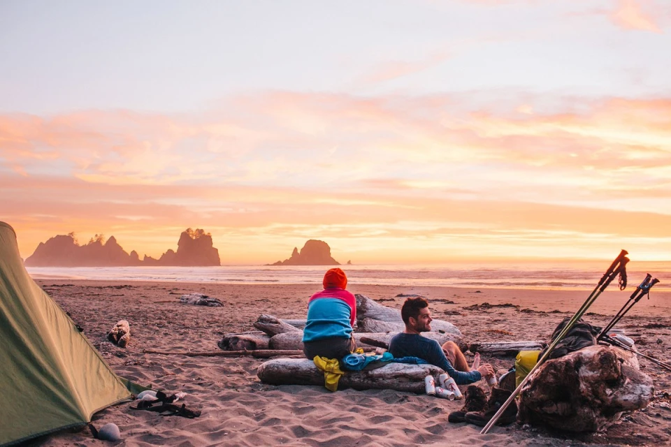 A couple sits on a log near a tent on a beach at sunset, with sea stacks in the background and two trekking poles nearby.