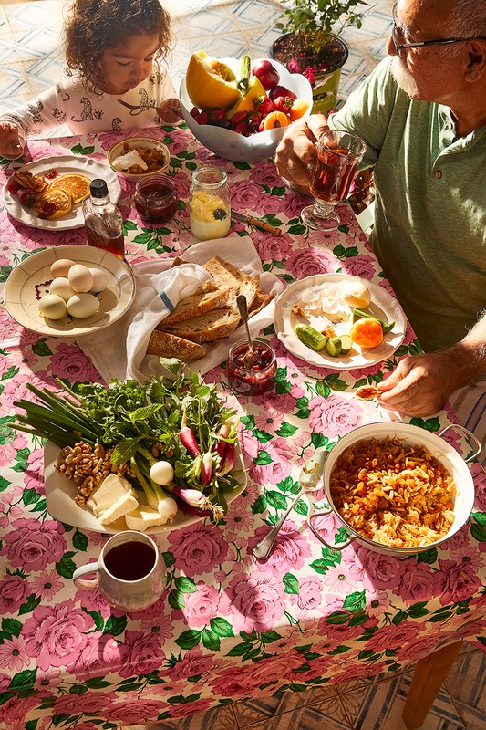 Overhead shot of a child and senior man at a breakfast table laden with various dishes like pancakes, fruit, bread, and cooked vegetables.