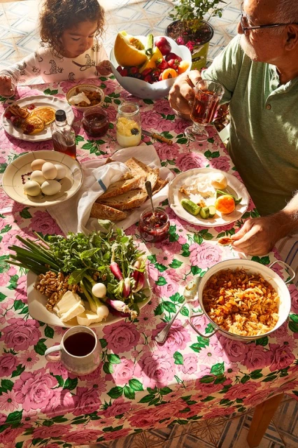 Overhead shot of a child and senior man at a breakfast table laden with various dishes like pancakes, fruit, bread, and cooked vegetables.