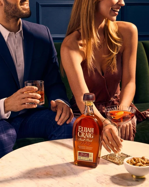 Elijah Craig bourbon bottle on a marble table with a man and a woman holding cocktails, and a bowl of nuts in the background.