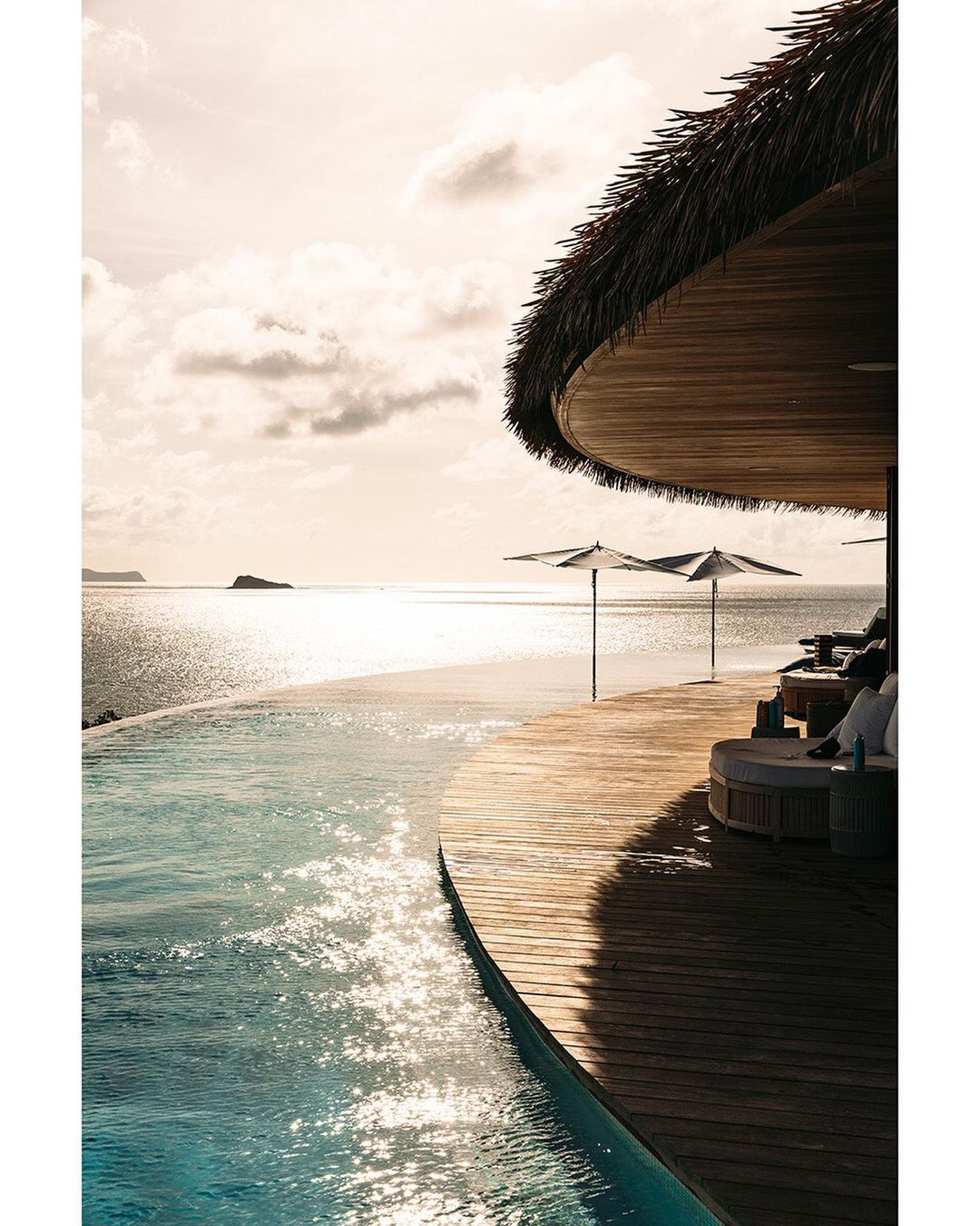 Eye-level shot showcases an infinity pool leading to the ocean, framed by a wooden terrace with sunbeds, umbrellas, and a thatched roof structure.