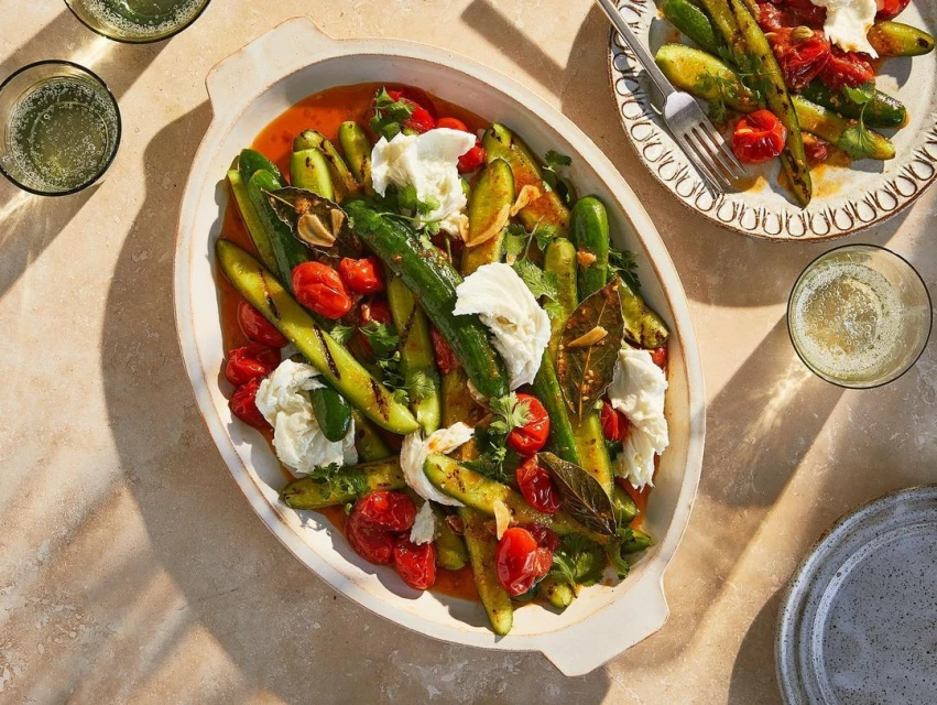 Overhead shot of grilled zucchini salad with burrata cheese, tomatoes, and herbs in a creamy dish with drinks and serving plates.