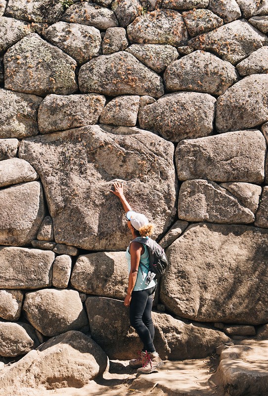Woman touches large stone wall during daytime. The wall is constructed of stones of various sizes. The woman is wearing a hat and backpack.