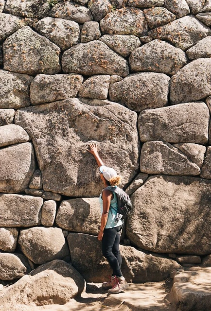 Woman touches large stone wall during daytime. The wall is constructed of stones of various sizes. The woman is wearing a hat and backpack.