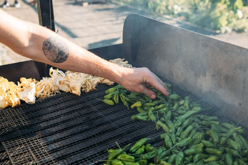 Close-up of a man's hand arranging green Shishito peppers and mushrooms on a metal grill for outdoor cooking.