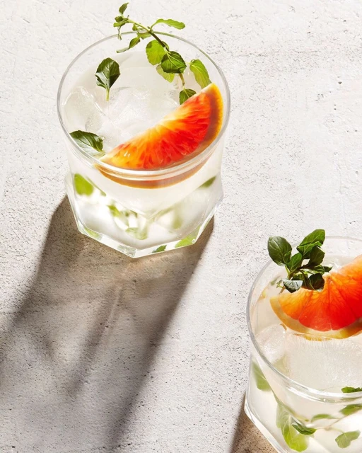 Two refreshing grapefruit cocktails in clear glasses with ice cubes and mint garnish on a textured white surface under bright light.