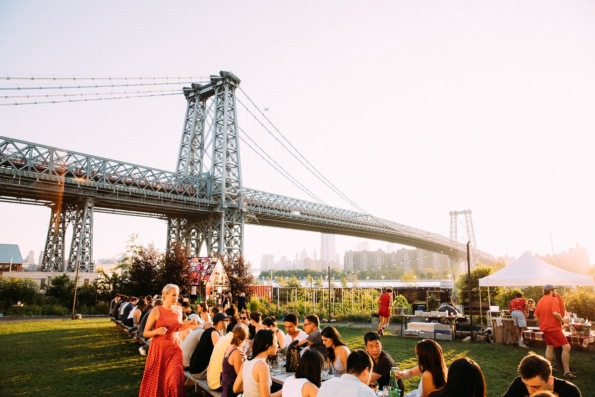 Outdoor gathering near Williamsburg Bridge with people at long tables on green grass, and distant city buildings in hazy sunlight.