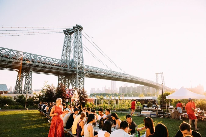 Outdoor gathering near Williamsburg Bridge with people at long tables on green grass, and distant city buildings in hazy sunlight.