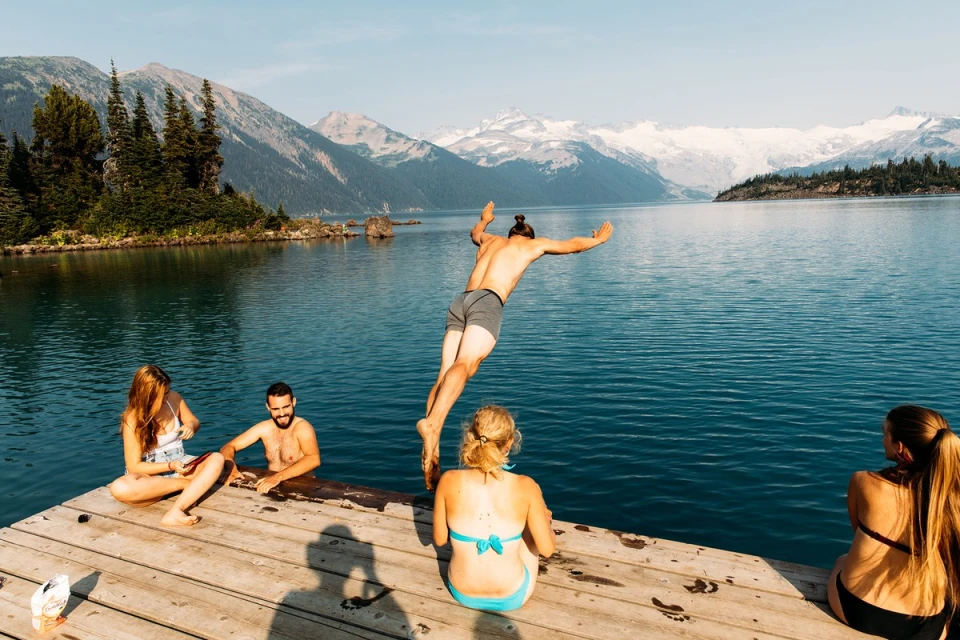 A group of people on a wooden dock next to a lake with one person diving into the water, with snow-capped mountains in the background on a sunny day.