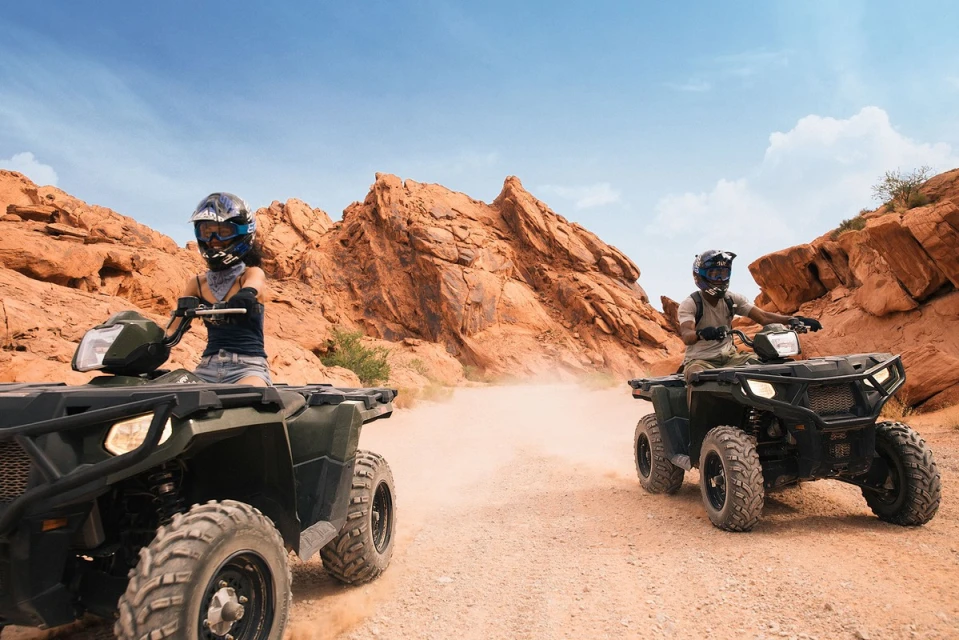 Two people riding ATVs on a sandy trail through a desert landscape with red rock formations under a clear blue sky.