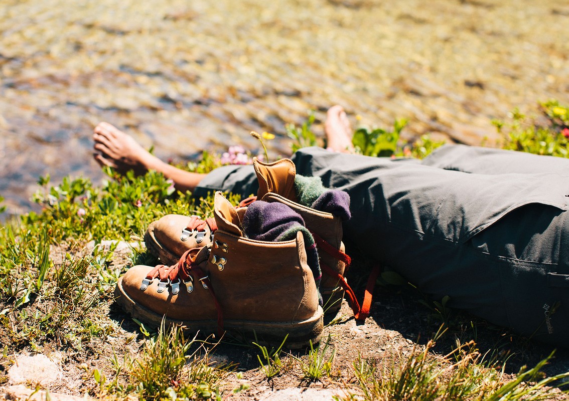 Hiking boots and person lying on the grass next to water. Brown leather boots with red laces and green grass in an outdoor nature setting.