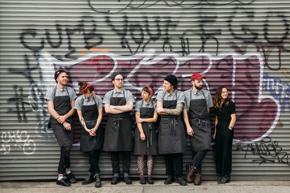 Group of seven people in aprons posing outdoors against a graffiti-covered corrugated metal wall, wearing casual clothing, in an urban setting.
