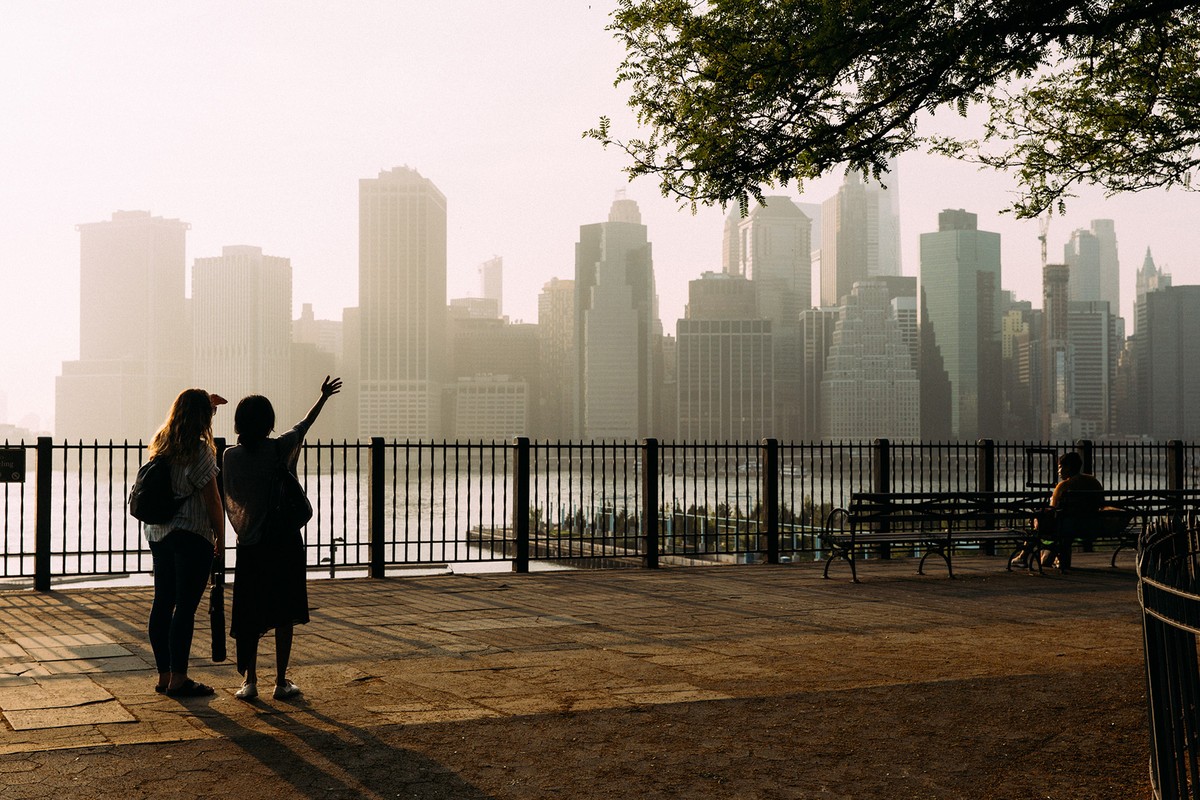 Two women on waterfront overlook with a cityscape view. Trees provide a partial frame around the skyline, fence separates walkway from water.