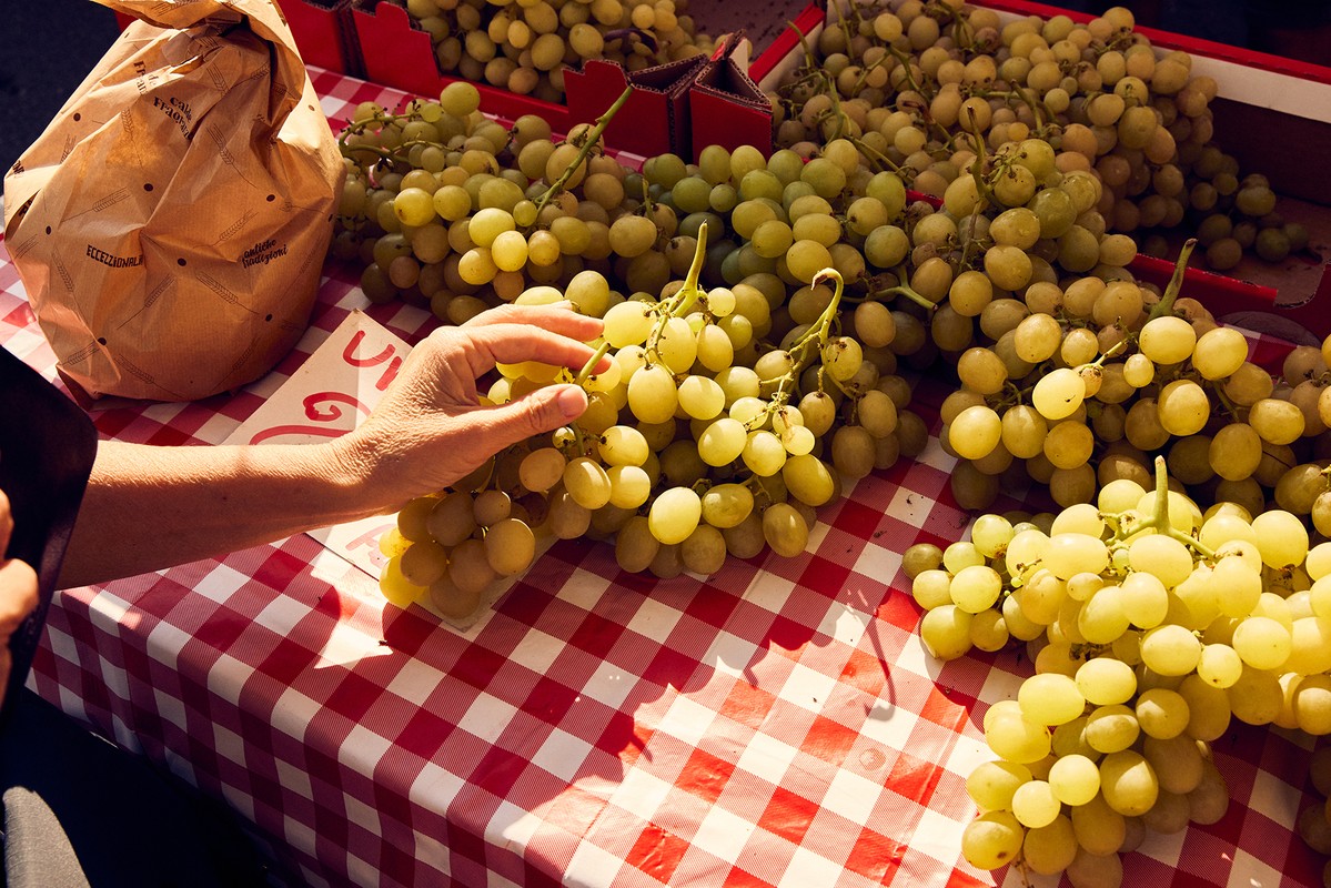 Close-up of a person's hand selecting green grapes on a red and white checkered tablecloth at a vibrant outdoor market.