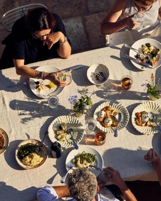 Overhead shot of an outdoor luncheon table with diners, plates of food, glassware, white tablecloth, and dappled sunlight.