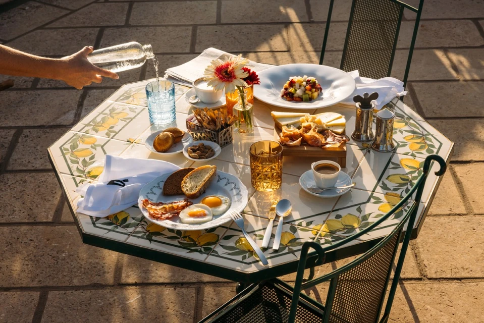 Outdoor breakfast table with water pouring into a glass, featuring fruit salad, cheese, toast, eggs, bacon, coffee, flowers, and tiled tabletop.