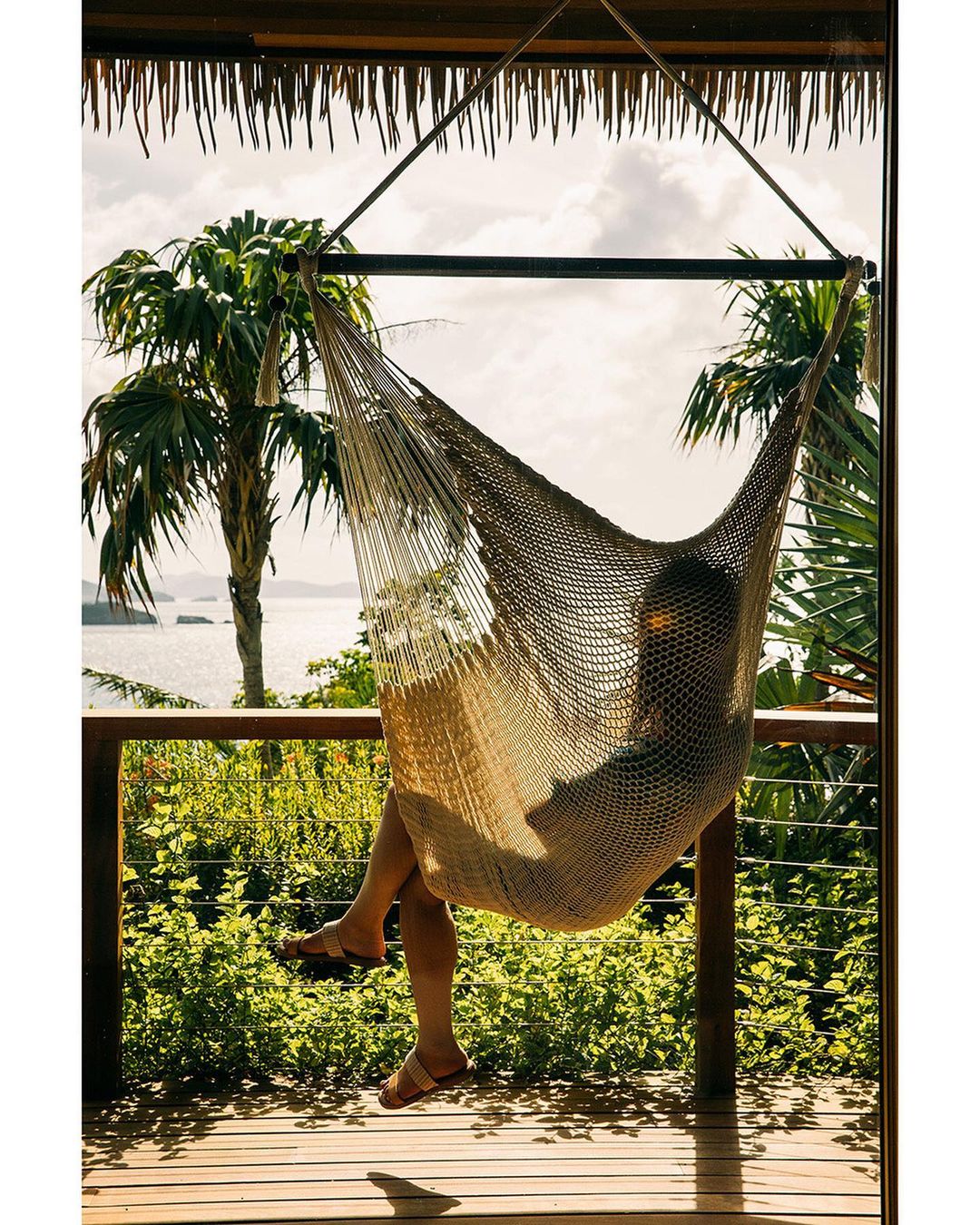 Adult relaxing in a net hammock suspended from a covered wooden patio with a view of a tropical shoreline, palm trees, and ocean.