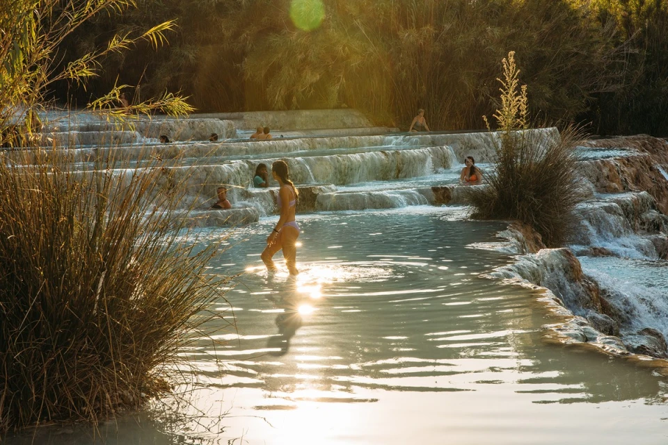 People relax in the tiered thermal pools of Cascate del Mulino, Italy, under a sunny sky with natural travertine formations and vegetation.