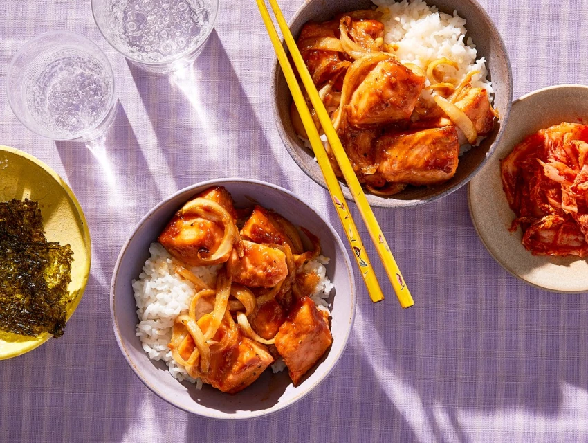 Overhead shot of two bowls of teriyaki salmon with rice and onions, kimchi, seaweed, chopsticks and glasses of water on lavender tablecloth.