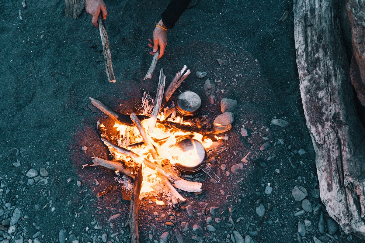 Campfire on a sandy beach. A person adds logs to the burning fire. Cooking pots are near the fire. Driftwood and rocks surround the fire.