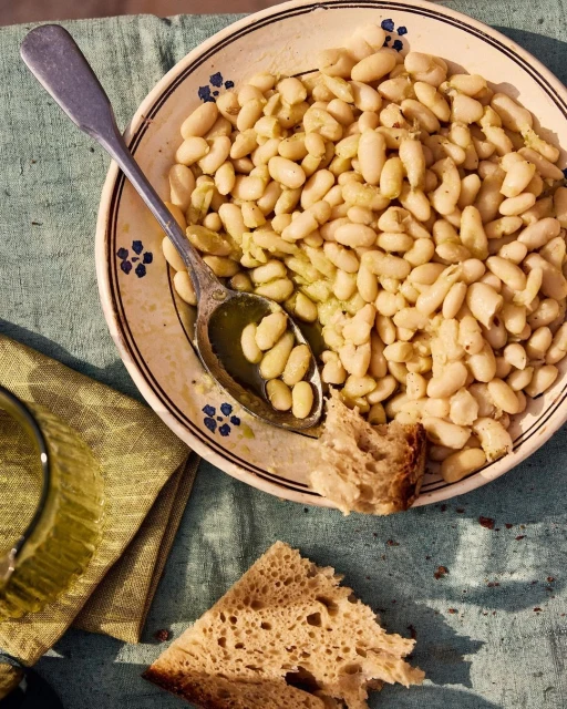 Overhead shot showcases a rustic plate filled with white beans, a silver spoon, pieces of crusty bread, and a glass on a textured surface.