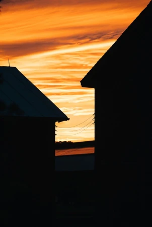 Silhouette of two buildings against an orange and yellow sky at sunset. Electrical wires run between the structures.