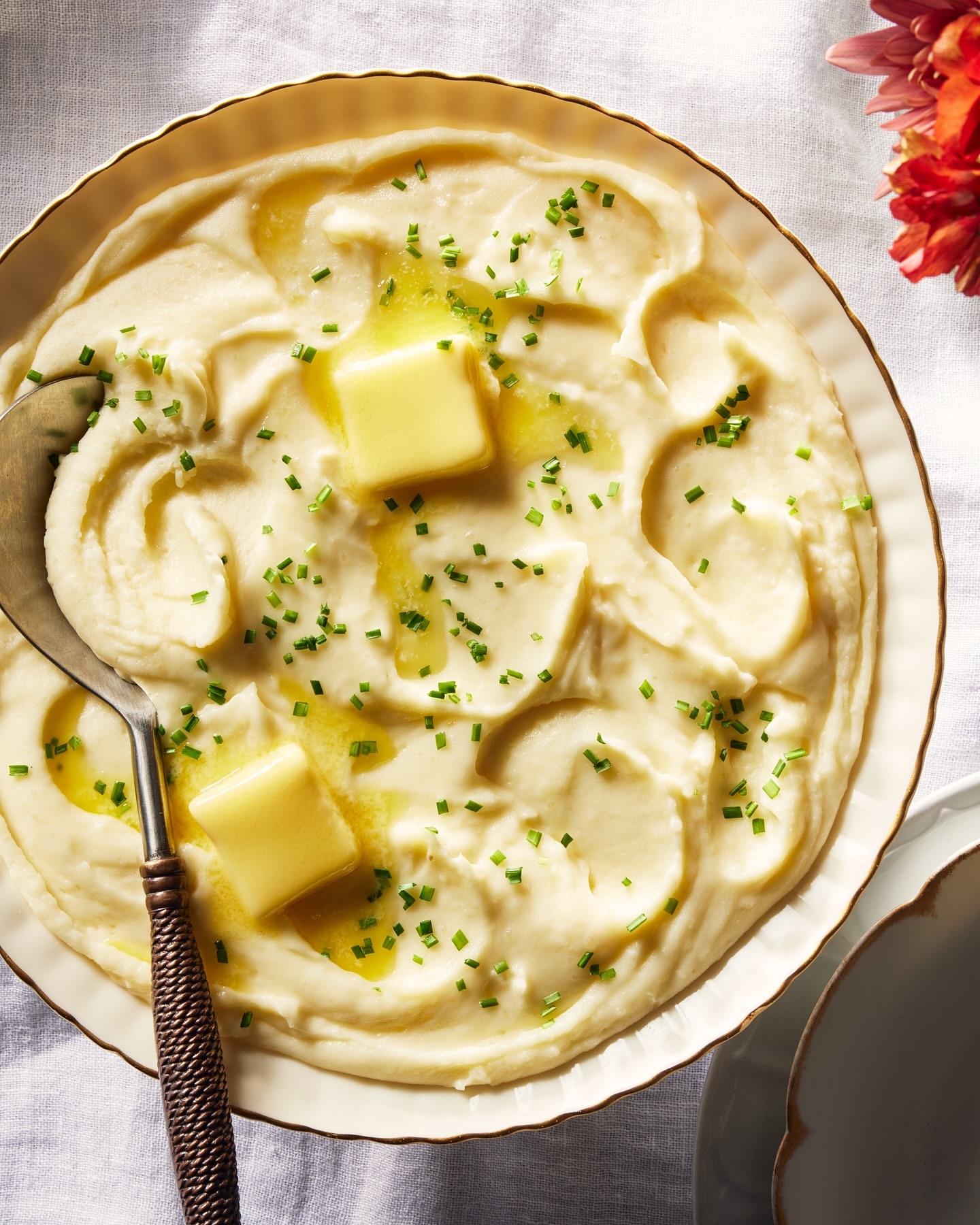 Overhead shot of creamy mashed potatoes in a white bowl, topped with butter cubes and chives, next to a metal spoon and floral decoration.