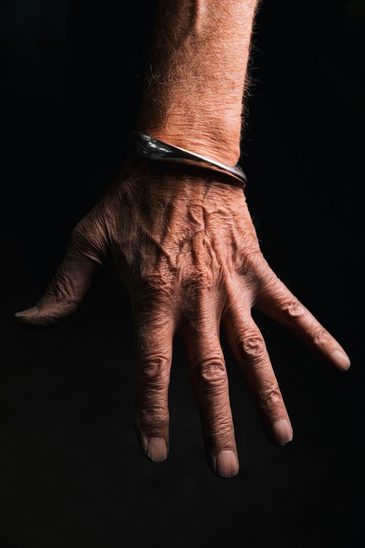 Close-up of an elderly adult's wrinkled hand with a metal bracelet against a dark background, showing the texture of skin with studio lighting.