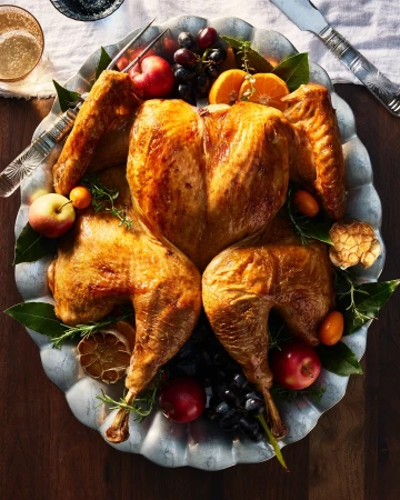 Roasted turkey centerpiece surrounded by fruit and herbs on silver platter, overhead shot showcasing golden-brown skin and festive presentation.