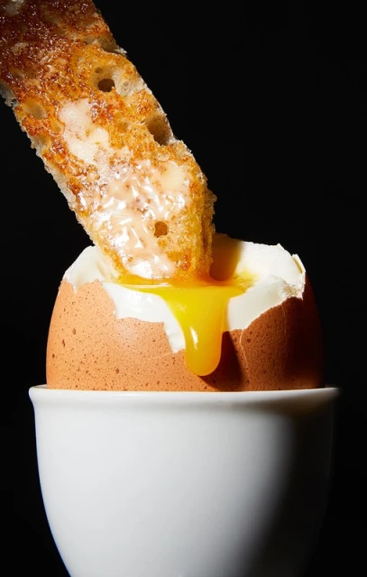 Runny egg in a white cup with toast being dipped in the yolk, set against a stark black background, food photography.