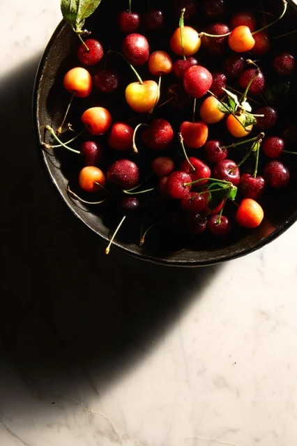 Overhead shot of fresh cherries, showcasing a mix of red and yellow varieties, presented in a dark bowl on a white marble surface.