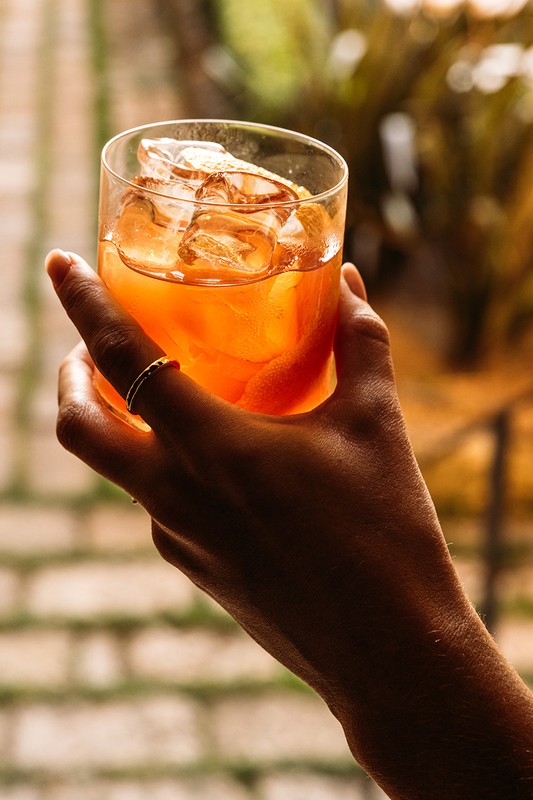 Close-up of a hand holding a glass of amber-colored cocktail with ice cubes and a citrus peel garnish, against a blurred background.