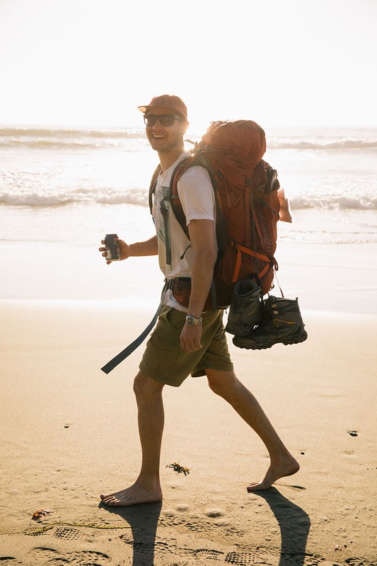 Man with backpack walking barefoot on beach. He wears shorts, t-shirt, baseball cap, sunglasses, and carries a beverage in his hand.