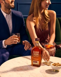 Man and woman seated together in a booth with a bottle of Elijah Craig bourbon and cocktails on a marble table.