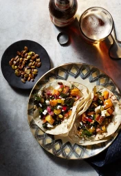 Overhead food photography of two filled tacos on a patterned plate with toasted seeds and a glass of beer.