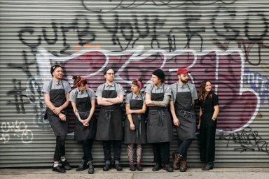 Group of six people in work aprons standing in a line in front of a metal roller shutter with graffiti