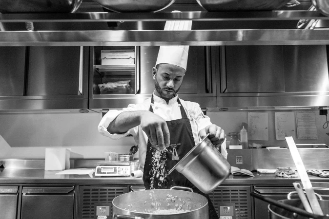 Monochrome photograph of a chef in a tall toque and apron seasoning a large pot in a stainless steel commercial kitchen.