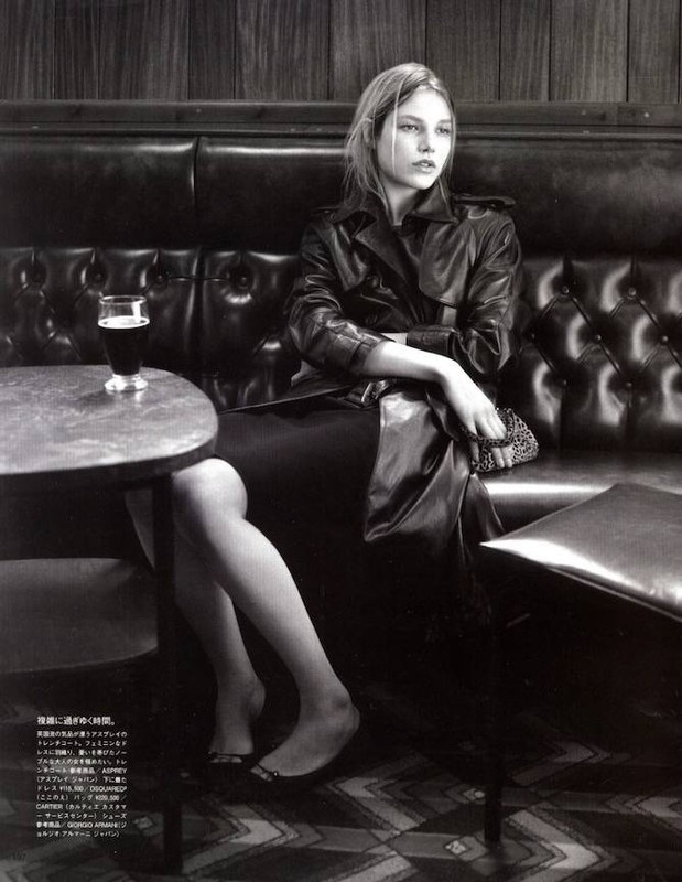 A monochrome fashion shot of a woman seated at a bar, wearing a trench coat, with a glass of dark beer on the table.