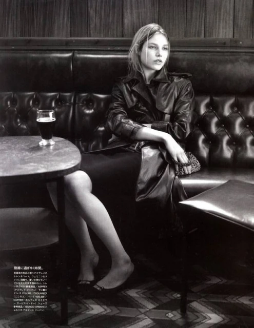 A monochrome fashion shot of a woman seated at a bar, wearing a trench coat, with a glass of dark beer on the table.