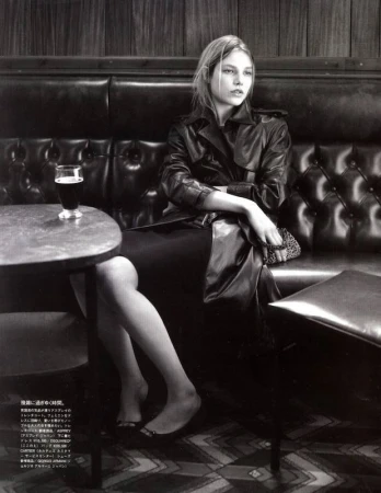 A monochrome fashion shot of a woman seated at a bar, wearing a trench coat, with a glass of dark beer on the table.
