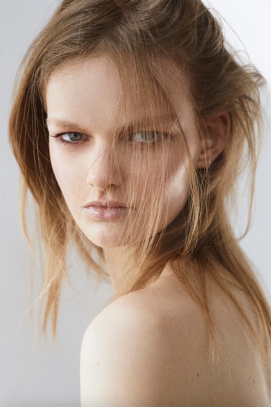 Close-up portrait of a woman with loose blonde hair falling across her face and minimalist makeup against a light background.