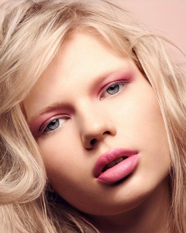 Close-up portrait of a woman with blonde hair and pink makeup. The model poses in front of a soft pink background in a beauty shot.