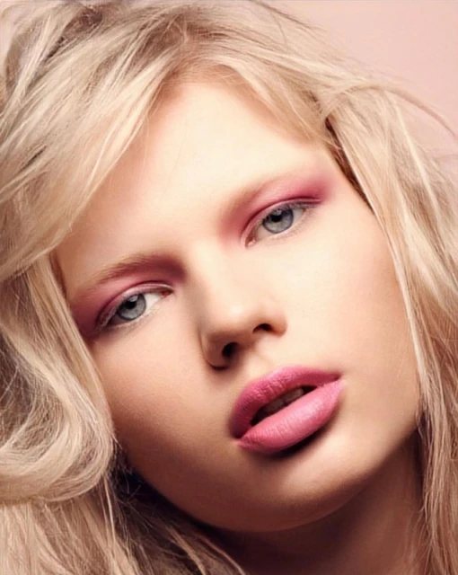 Close-up portrait of a woman with blonde hair and pink makeup. The model poses in front of a soft pink background in a beauty shot.