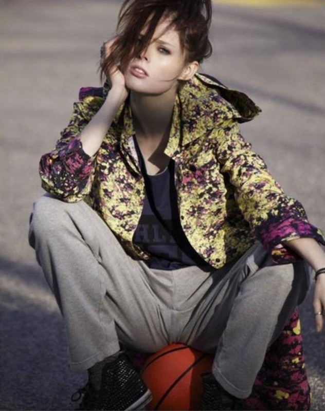 Model squatting outdoors while resting on a basketball, wearing a patterned yellow jacket and grey sweatpants.