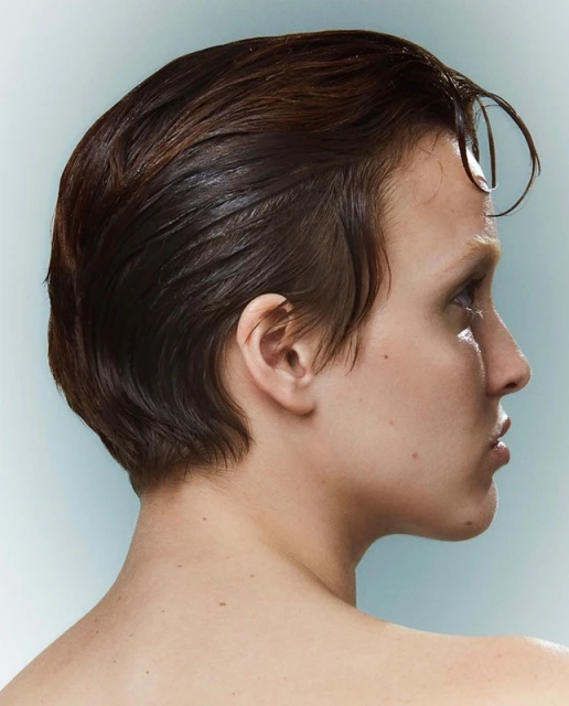 Close-up side profile of a young adult with wet brunette hair, fair skin, bare shoulder, and a light blue-grey background.